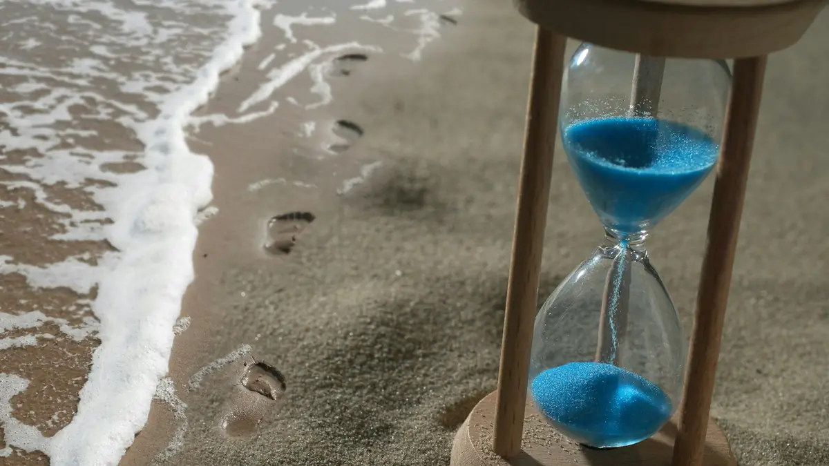 Wooden hourglass with blue sand standing on a wet beach beside footprints
