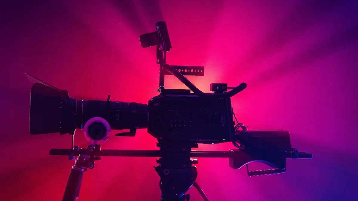 Cinema camera on a tripod silhouetted against pink and purple light beams