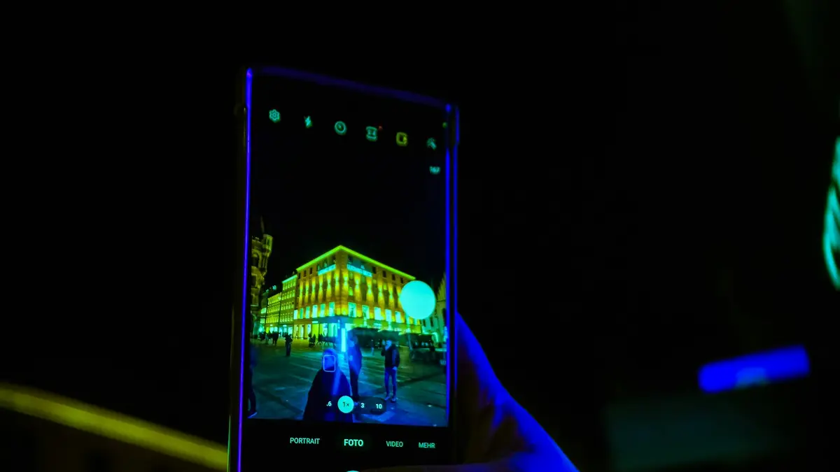 Hand holding a smartphone in the dark, its camera screen showing a building lit in neon green and yellow