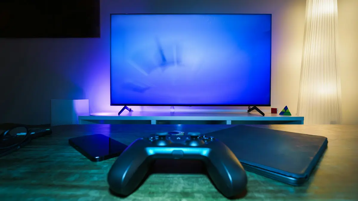 Black game controller on a wooden table in front of a TV glowing blue in a dim living room