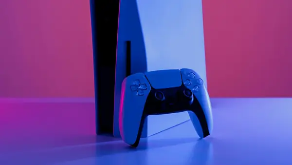 White game console standing upright next to its controller in blue and pink light