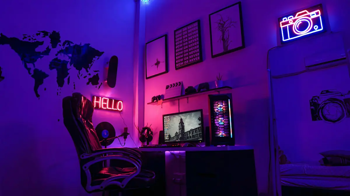 Gaming desk in a dark room lit pink and purple, with a monitor, a PC tower and neon signs on the walls