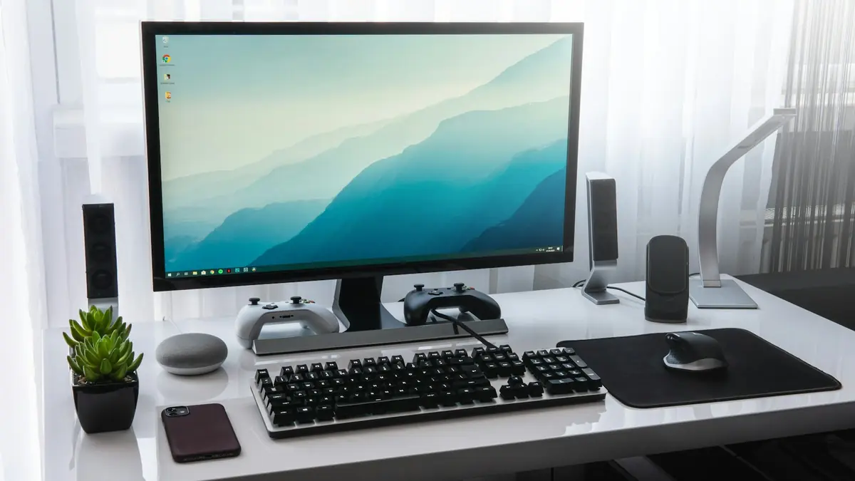 Desktop PC setup on a white desk with a monitor, keyboard, mouse, speakers and two game controllers